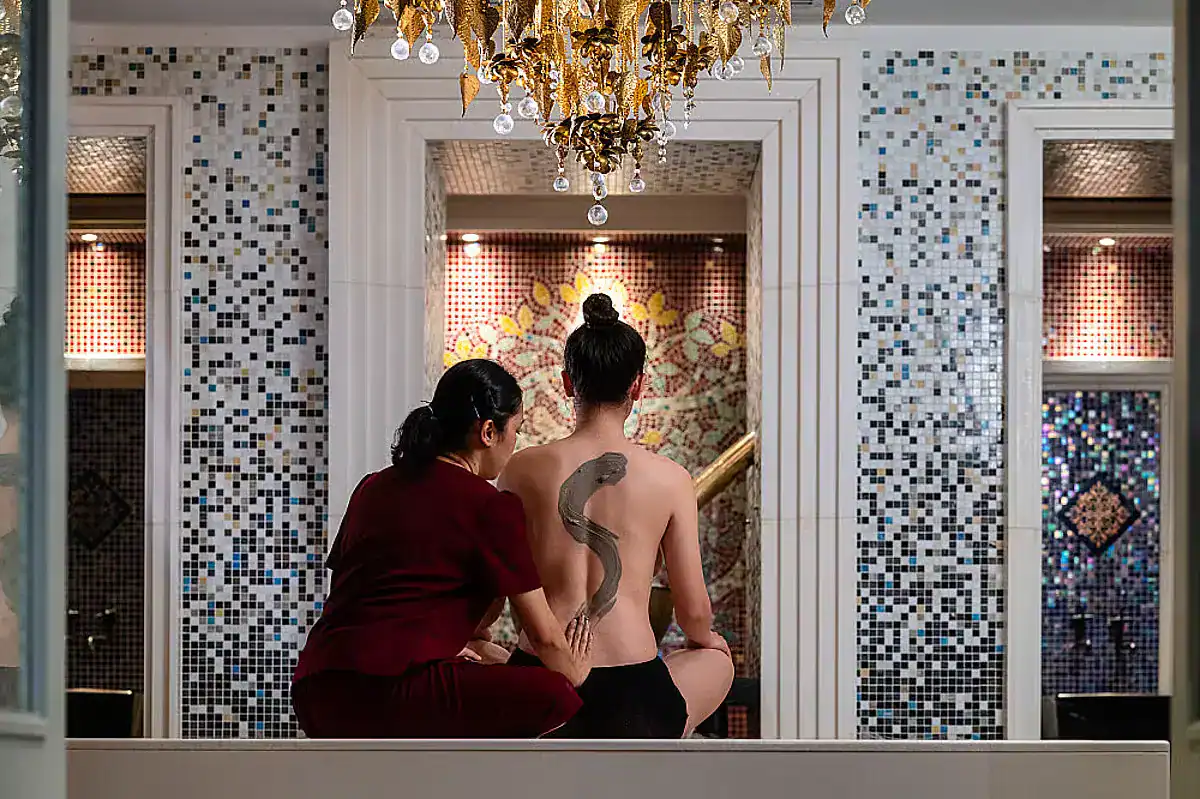 A woman has her back turned to a staff member, who gently rubs healing oils and clay onto her skin. They are in a luxurious room filled with granite walls and a gold chandelier.