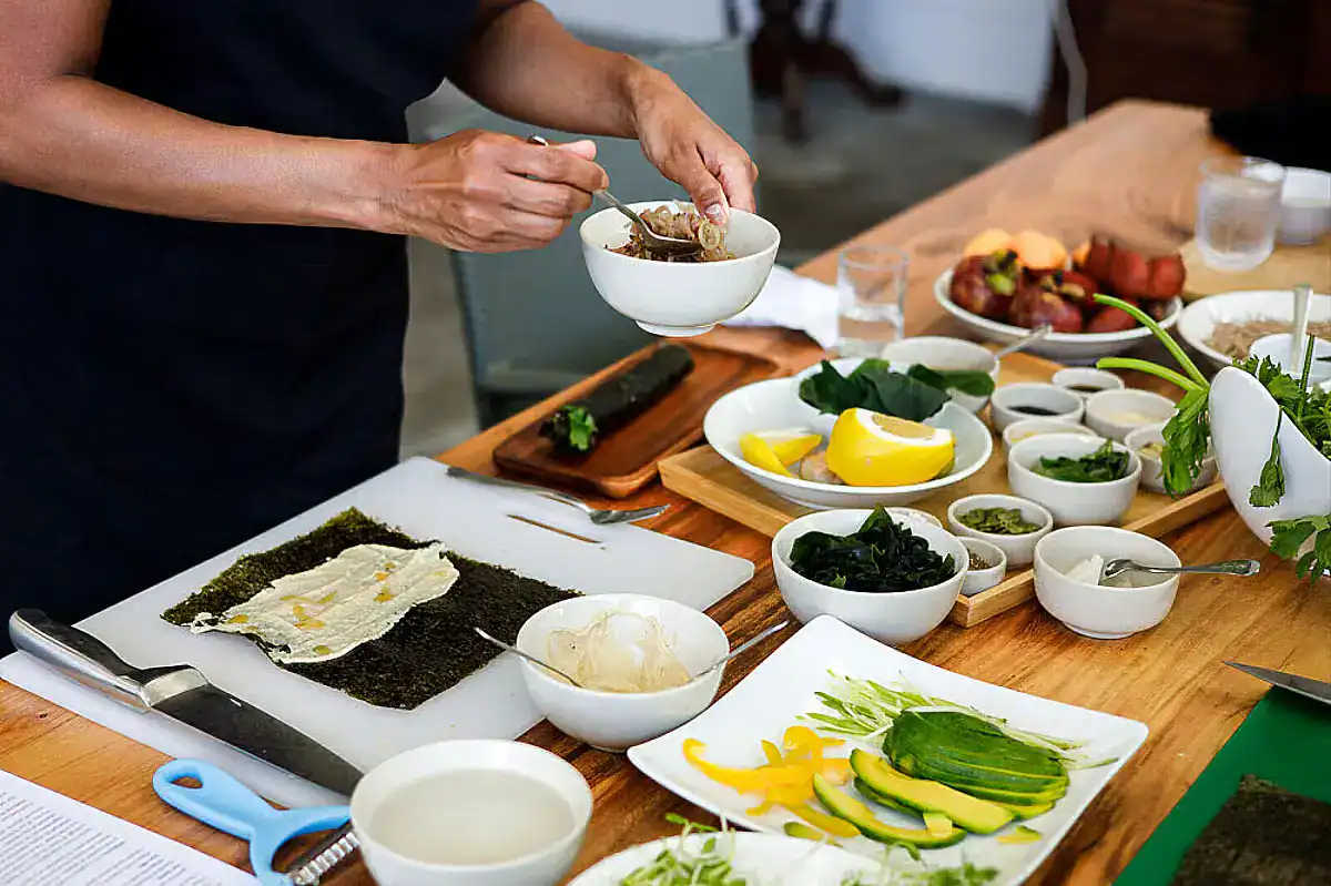 A long wooden table filled with plates and bowls of food, mainly vegetable-forward meals, at CC's Hideaway, one of the best detox retreats in Phuket. The hands of a person, presumably the chef, can be seen plating one of the bowls.