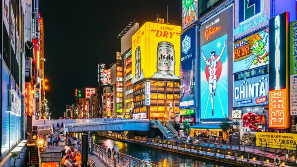 Night view of Dotonbori in Osaka with bright neon signs, the Glico running man billboard, and crowds along the canal.