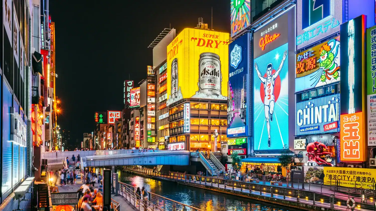 Night view of Dotonbori in Osaka with bright neon signs, the Glico running man billboard, and crowds along the canal.