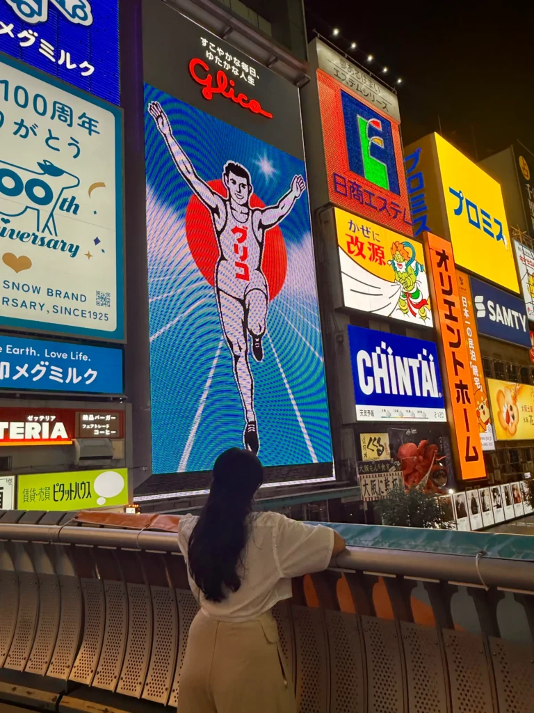 Things to Do in Osaka at Dotonbori featuring the Glico Man billboard glowing at night with neon signs and a visitor watching from the bridge.