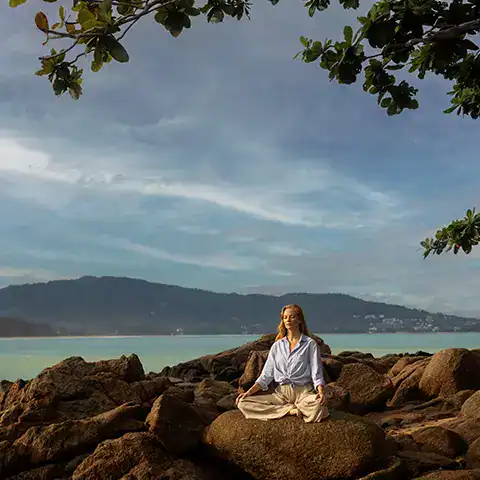 A person sits in cross-legged on the rocky facing the ocean. They appear to be meditating alone.