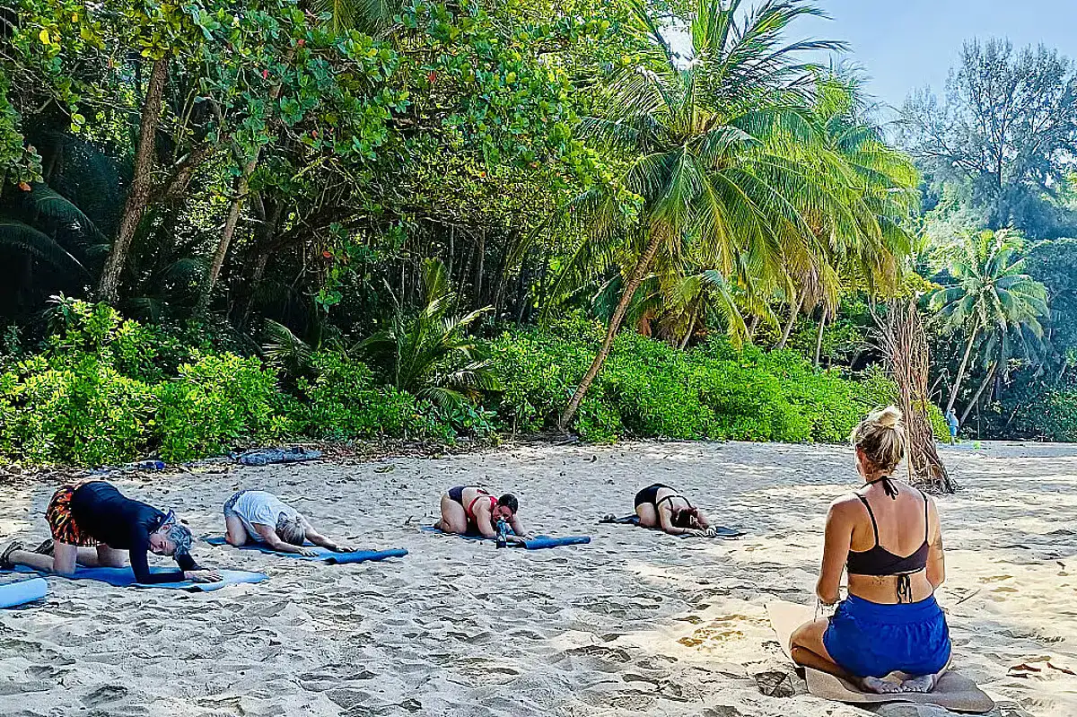 Participants of The Yoga Lodge, one of the most calming wellness retreats in Phuket, lie on their yoga mats while a female instructor watches over them. They are doing their exercise on the sand, framed by the greenery of Surin beach.