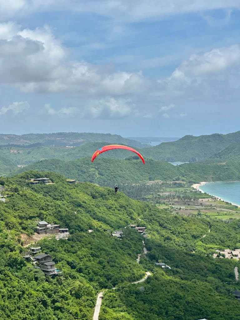 Paraglider flying over green hills and coastline in Lombok, a memorable adventure and one of the most unique things to do in Kuta Lombok