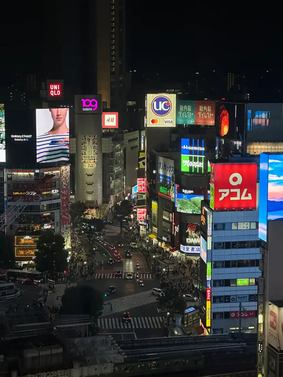 A nighttime view of Shibuya intersection filled with cars and crowds surrounded by tall buildings covered in bright illuminated billboards.