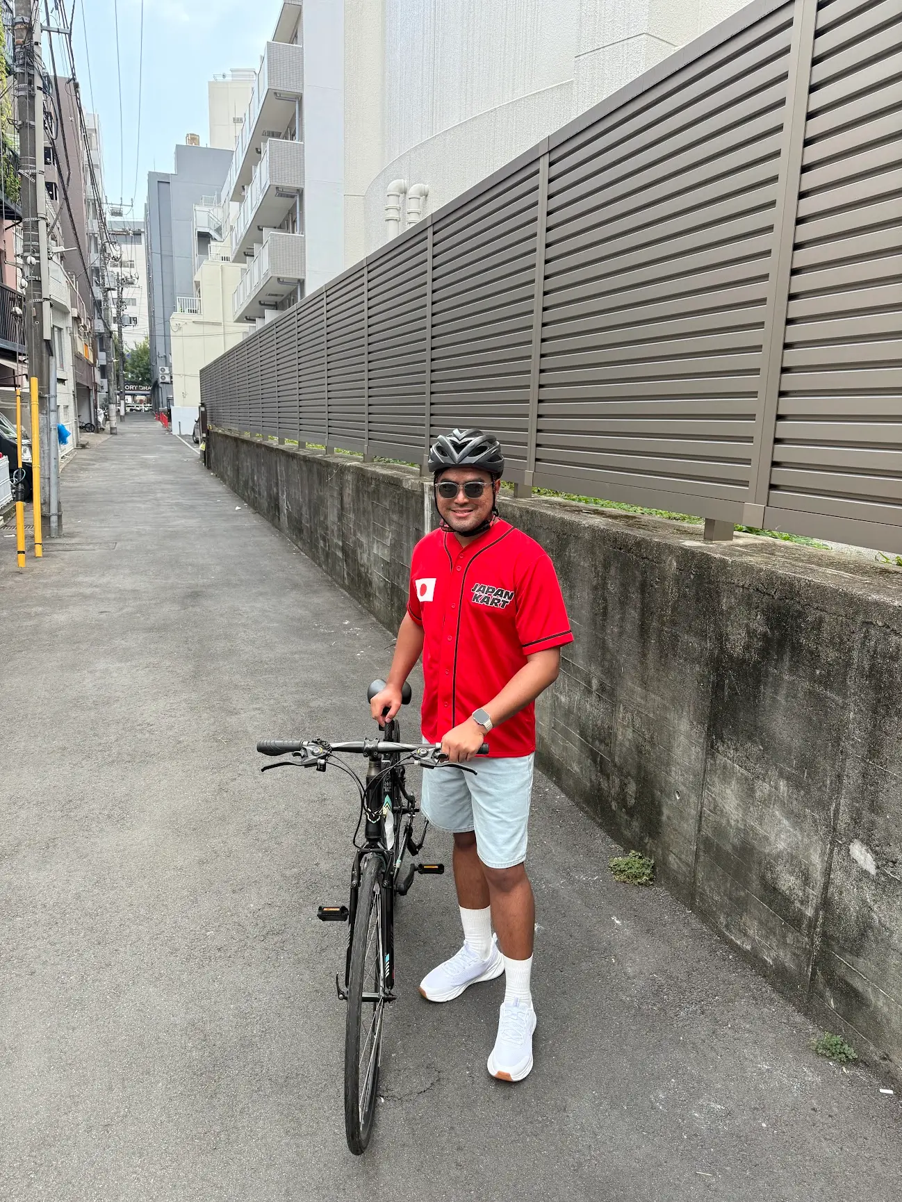 A man wearing a red Japan Kart shirt, helmet, and sunglasses stands with a bicycle on a quiet street in Japan.