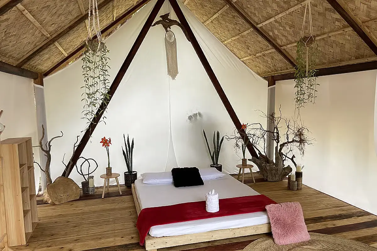 A bed with complimentary towels, a desk, and some potted plants in a shaded nipa hut.