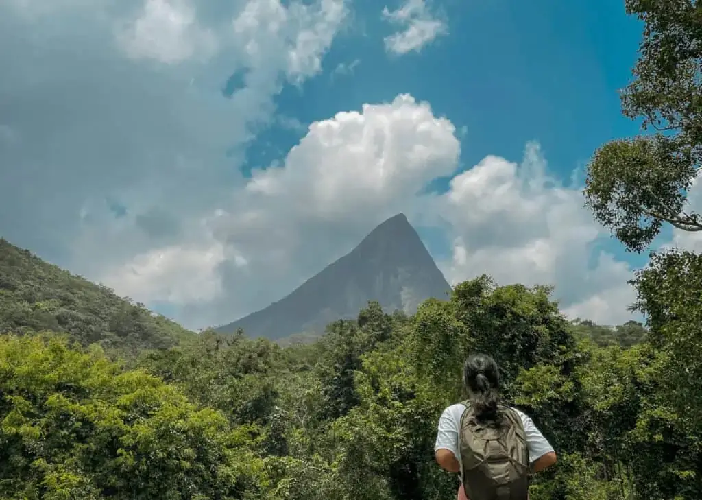 A woman carrying a backpack is facing a lush green mountains, likely on the way to the yoga retreats in Sri Lanka.