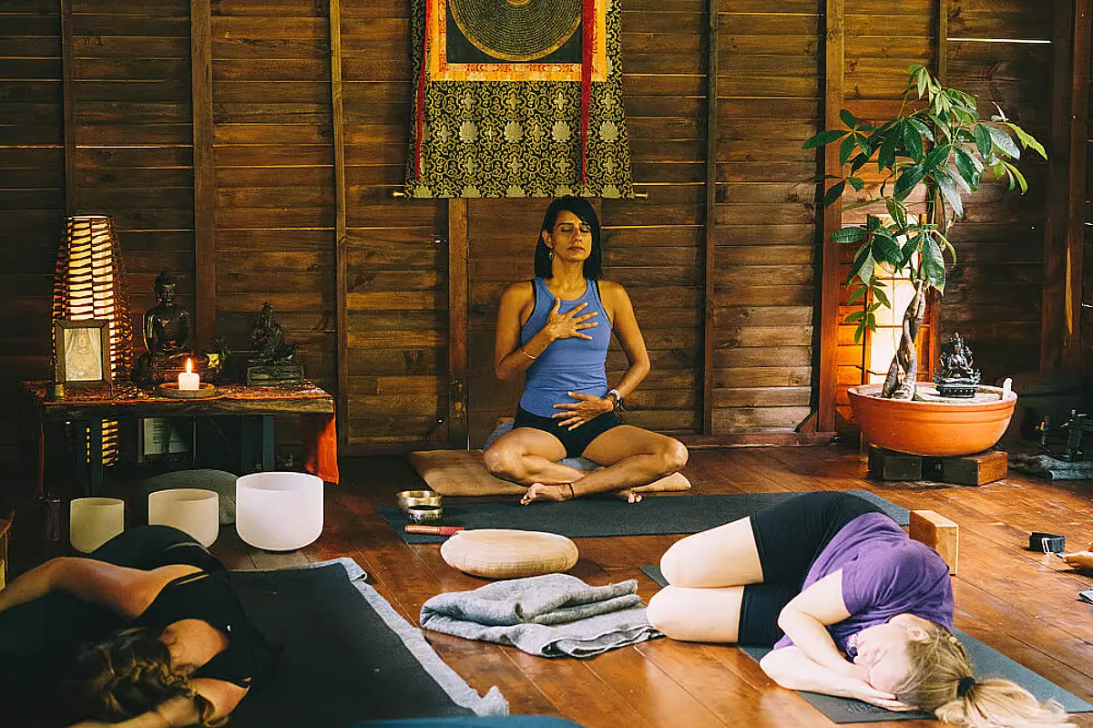 A woman sits cross leg on a yoga mat, meditating, while two others lie on their side.