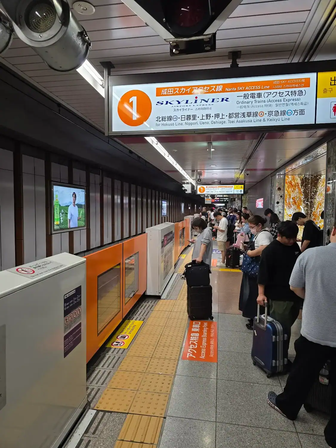 Passengers waiting with luggage on a platform at Narita Airport Station in Japan, under signs for the Keisei Skyliner and Sky Access Line.