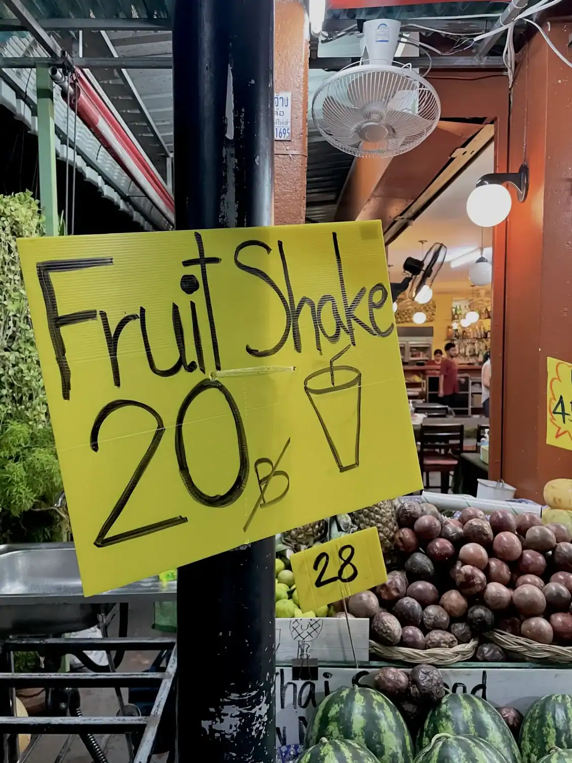 Bright yellow sign reading “Fruit Shake 20฿” displayed beside fresh passion fruit and watermelons at a street fruit stall in Phuket.