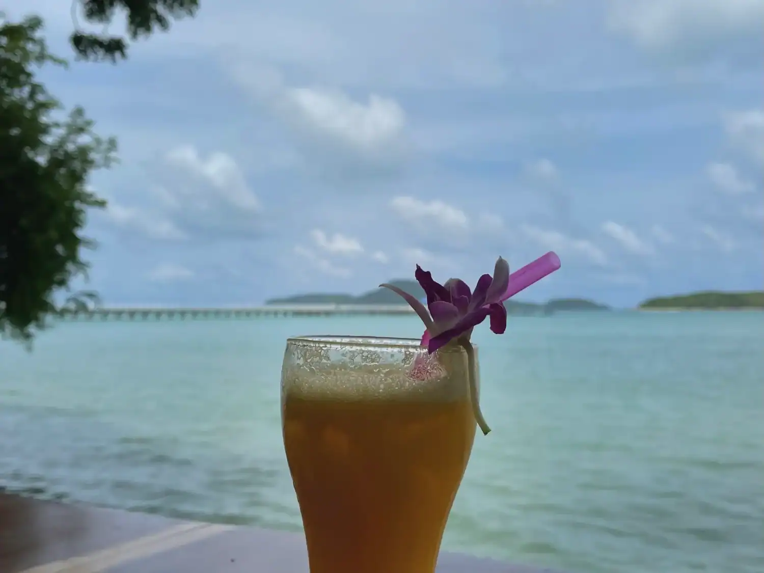 Mango shake topped with a purple orchid and pink straw at Salaloy Seafood Restaurant in Rawai. The tropical drink sits beside the ocean with cloudy blue skies and distant islands in the background, highlighting a scenic spot for where to eat in Phuket.