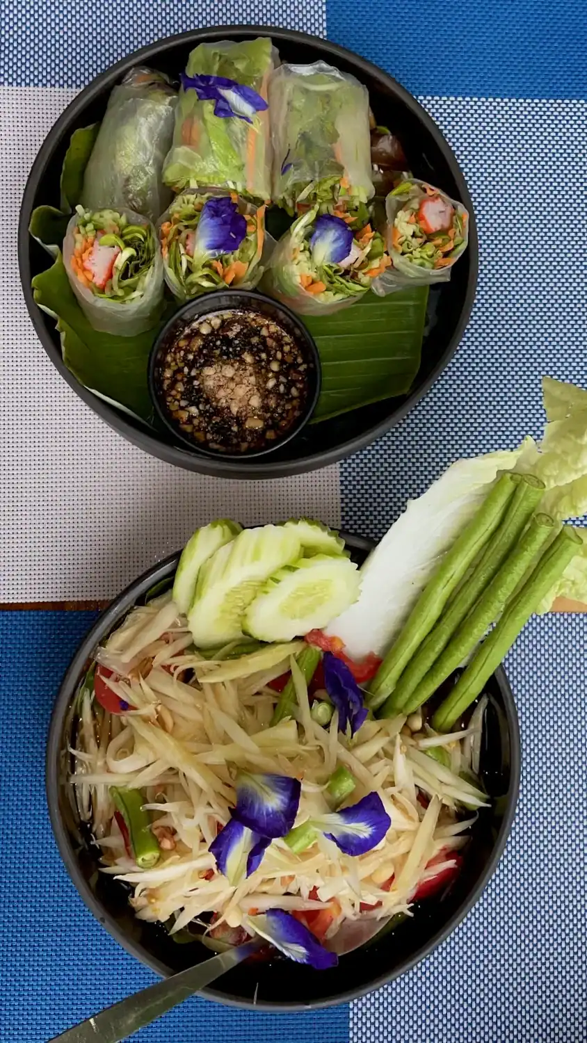 Fresh Thai spring rolls filled with vegetables and crab sticks served with dipping sauce beside a bowl of green papaya salad topped with edible blue flowers.