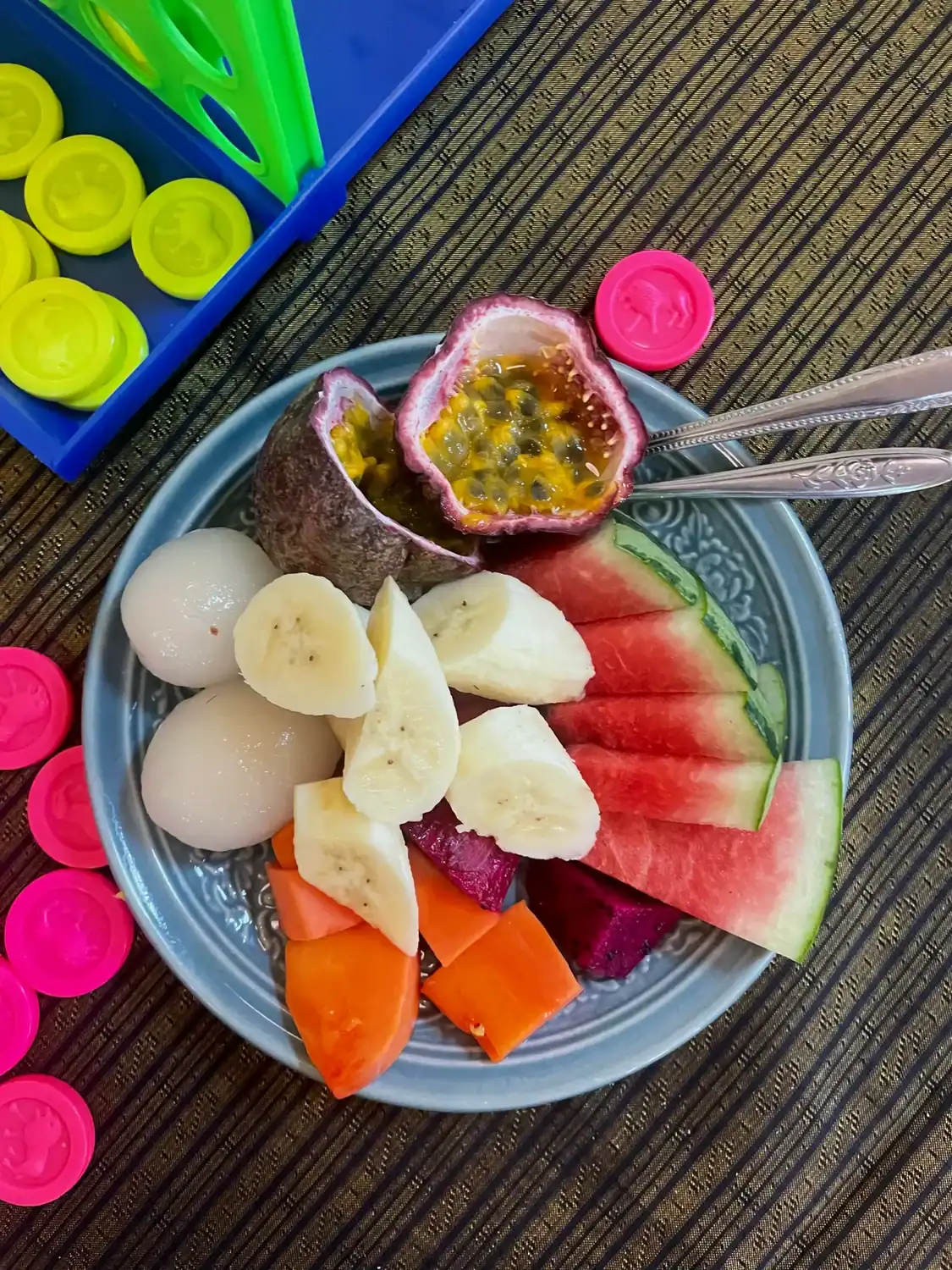 Plate of tropical fruit including watermelon, banana, papaya, dragon fruit, lychee, and fresh passion fruit served on a woven table mat in Phuket.