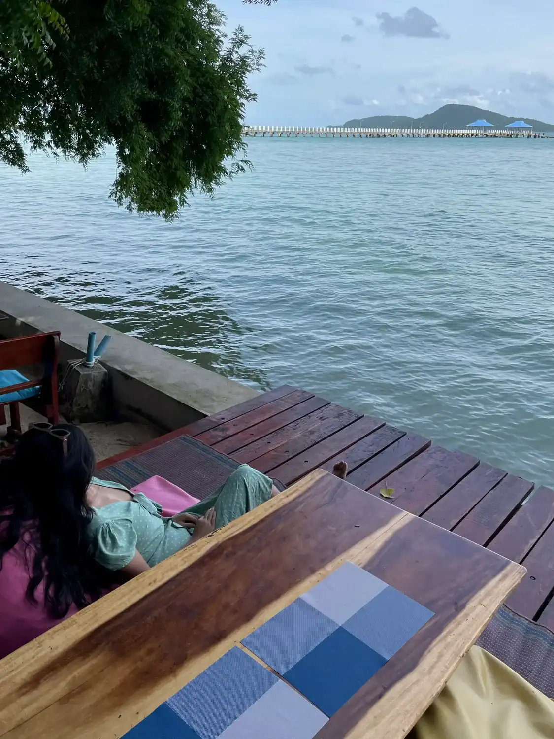 Wooden waterfront seating area overlooking the ocean in Rawai with calm water, cloudy skies, and a long pier stretching toward distant hills.