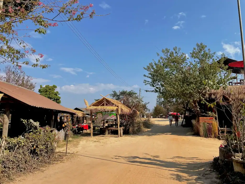 Volunteer in Thailand at a rural village community with local shops, traditional bamboo structures, and a quiet countryside road under a clear blue sky.