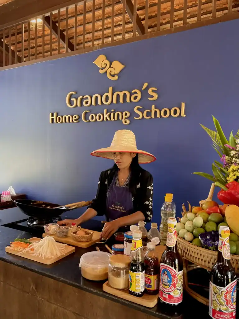 Thai cooking instructor demonstrating traditional recipes during a Chiang Mai cooking class at Grandma's Home Cooking School, surrounded by fresh local ingredients and cooking stations.