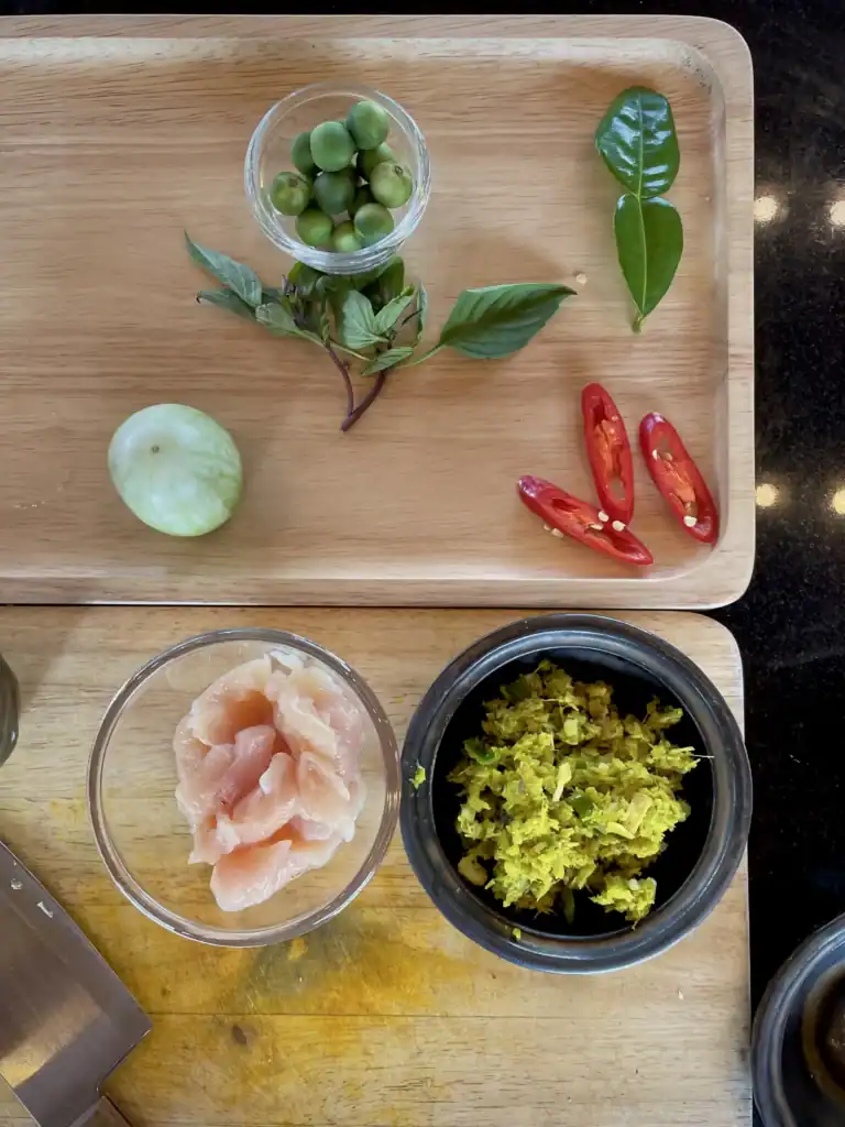 Fresh ingredients for a Chiang Mai cooking class, including Thai eggplant, Thai basil, kaffir lime leaves, red chilies, green curry paste, and sliced chicken arranged on a wooden tray before making authentic Thai green curry.