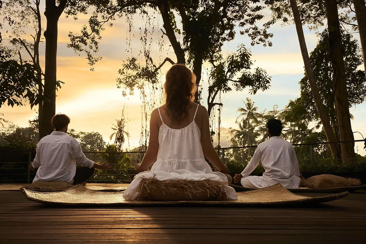 Three people meditating on a wooden deck at sunset at Ayurvedagram in Ubud, Bali, with jungle trees behind them