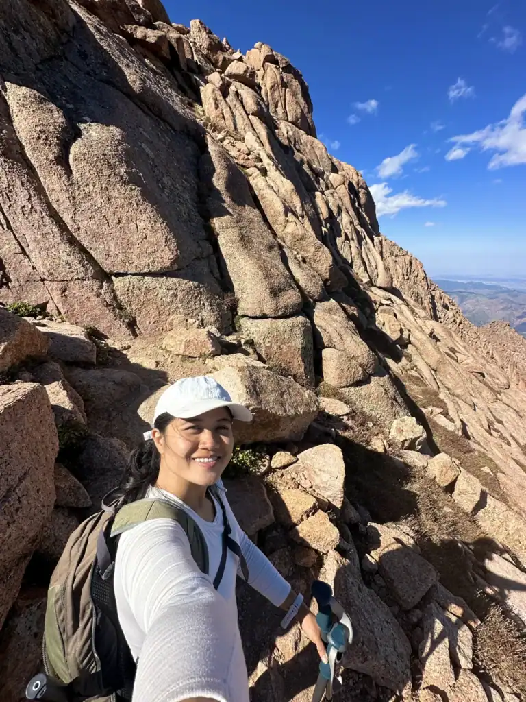 Me smiling on a rocky granite trail on Pikes Peak in Colorado, wearing a white cap and backpack and holding trekking poles