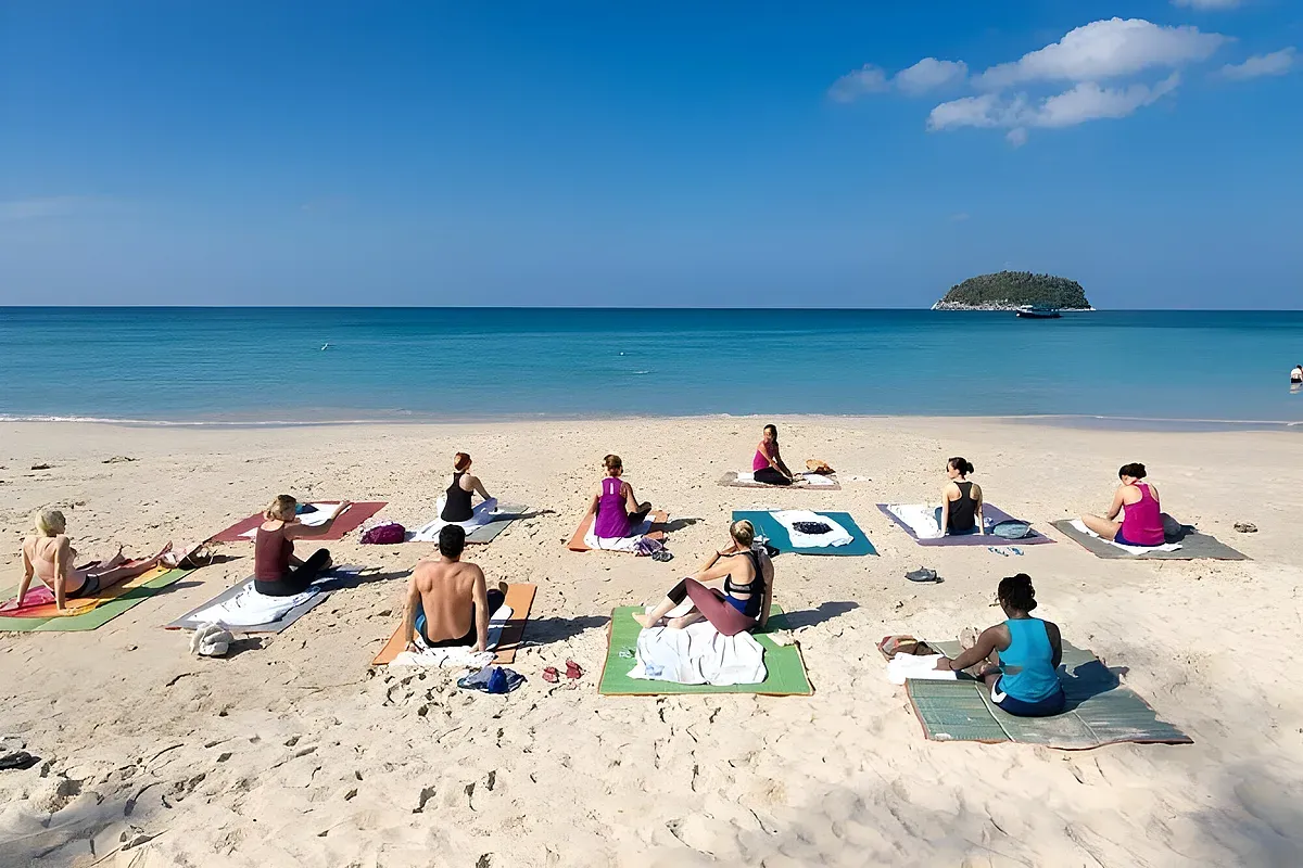 Small group practicing yoga on mats on a sandy Phuket beach with a small island in the distance