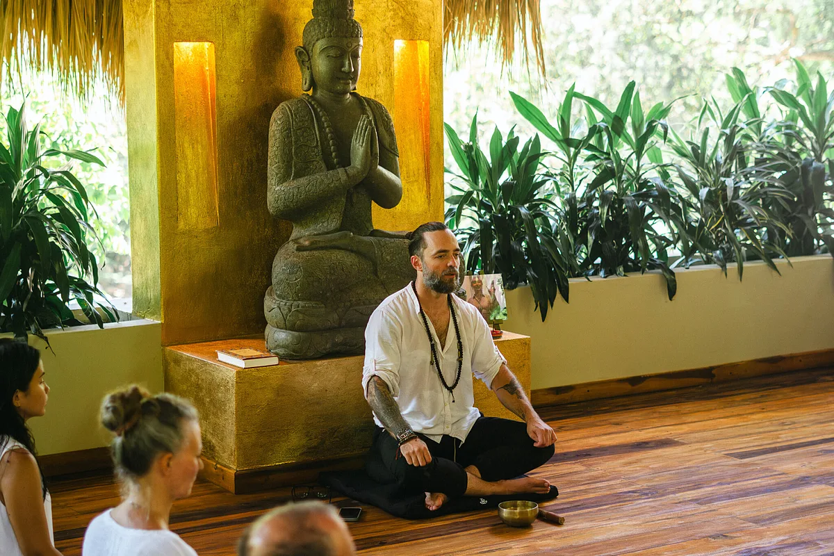 A teacher sitting in meditation in front of a Buddha statue with students beside him at the Senda Loto silent Buddhist retreat in Colombia
