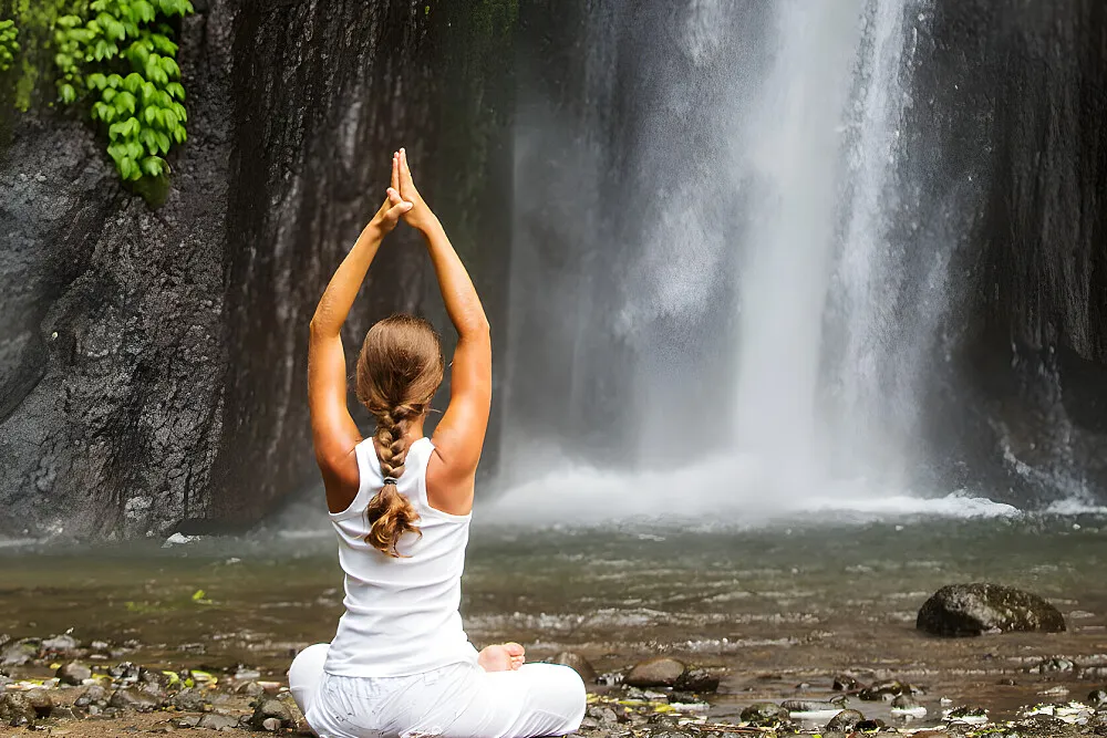 A woman in white sits in a prayer pose facing a waterfall